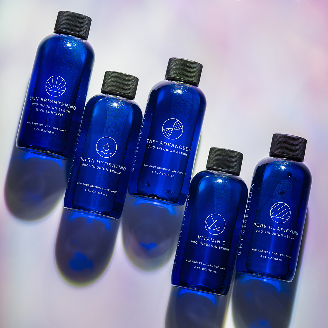 Image of DiamondGlow's SkinMedica Pro Infusion Serums' blue bottles flat laid on colorful background, with Skin Brightening, Hydrating, TNS Advanced+ Anti-Aging, Vitamin C, and Pore Clarifying from left to right