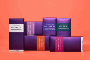 Juvederm collection of dermal hyaluronic acid fillers display of boxes, clockwise from left: Juvederm Voluma XC, Juvederm Ultra Plus XC, Juvederm Vollure XC, Juvederm Volux XC, Juvederm Volbella XC, and Juvederm Ultra XC on orange background